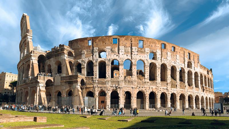The Colosseum in Rome under a bright sky, its arched tiers seen from the grass of the surrounding park