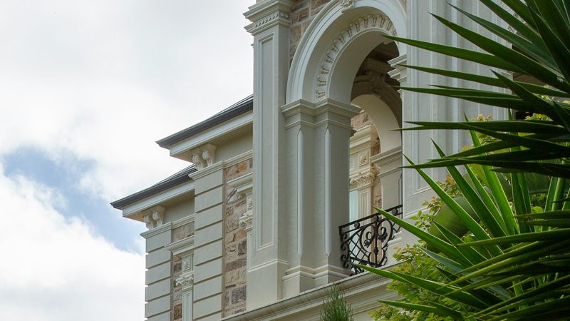 A large rendered and sandstone house with an arched loggia and wrought iron balcony, seen past palm fronds
