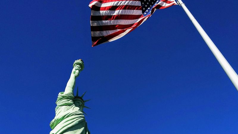 The Statue of Liberty raising her torch against a deep blue sky, the United States flag flying from a pole beside her